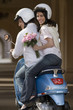 Photo: Couple riding on motor scooter near colonnade, woman holding bouquet of pink flowers, looking over shoulder, smiling, rear view, portrait Couple riding on motor scooter near colonnade, woman holding bouquet of pink flowers, looking over shoulder, smiling, rear view, portrait