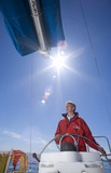 Photo: Man in red jacket standing at helm of sailing boat out at sea, steering (lens flare) Man in red jacket standing at helm of sailing boat out at sea, steering (lens flare)