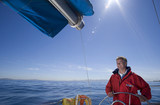 Photo: Man in red jacket standing at helm of sailing boat out at sea, smiling, steering (lens flare) Man in red jacket standing at helm of sailing boat out at sea, smiling, steering (lens flare)