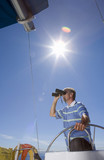 Photo: Man in striped blue polo shirt standing at helm of sailing boat out at sea, looking through binoculars, low angle view (lens flare) Man in striped blue polo shirt standing at helm of sailing boat out at sea, looking through binoculars, low angle view (lens flare)