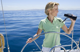 Photo: Woman in green polo shirt standing at helm of sailing boat out at sea, steering, looking through binoculars, smiling Woman in green polo shirt standing at helm of sailing boat out at sea, steering, looking through binoculars, smiling