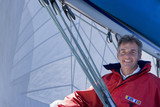 Photo: Man in red jacket sitting on deck of sailing boat below sail, smiling, portrait, low angle view Man in red jacket sitting on deck of sailing boat below sail, smiling, portrait, low angle view