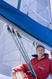 Photo: Man in red jacket sitting on deck of sailing boat below sail, smiling, low angle view Man in red jacket sitting on deck of sailing boat below sail, smiling, low angle view