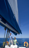 Photo: Couple standing at helm of sailing boat out to sea, woman steering, man leaning on sail rigging, smiling Couple standing at helm of sailing boat out to sea, woman steering, man leaning on sail rigging, smiling