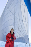 Photo: Woman in red jacket standing on deck of sailing boat below sail, holding binoculars, leaning on mast, smiling, front view, portrait Woman in red jacket standing on deck of sailing boat below sail, holding binoculars, leaning on mast, smiling, front view, portrait