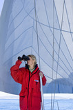 Photo: Woman in red jacket standing on deck of sailing boat below sail, looking through binoculars, leaning on mast Woman in red jacket standing on deck of sailing boat below sail, looking through binoculars, leaning on mast