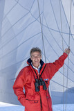 Photo: Man in red jacket standing on deck of sailing boat below sail, leaning on rigging, binoculars hanging around neck, smiling, front view, portrait Man in red jacket standing on deck of sailing boat below sail, leaning on rigging, binoculars hanging around neck, smiling, front view, portrait
