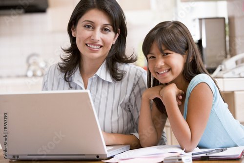 Photo: Woman and young girl in kitchen with laptop and paperwork smilin Woman and young girl in kitchen with laptop and paperwork smilin