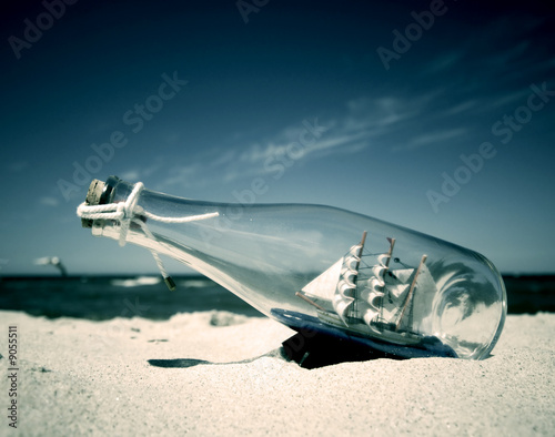 Photo: Bottle with ship inside lying on the beach. Conceptual image Bottle with ship inside lying on the beach. Conceptual image