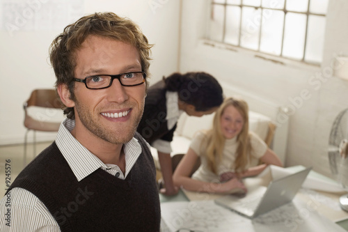 Photo: Young man smiling, women with laptop in background Young man smiling, women with laptop in background