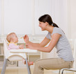 Foto: Mother feeding hungry baby in highchair in kitchen Mother feeding hungry baby in highchair in kitchen