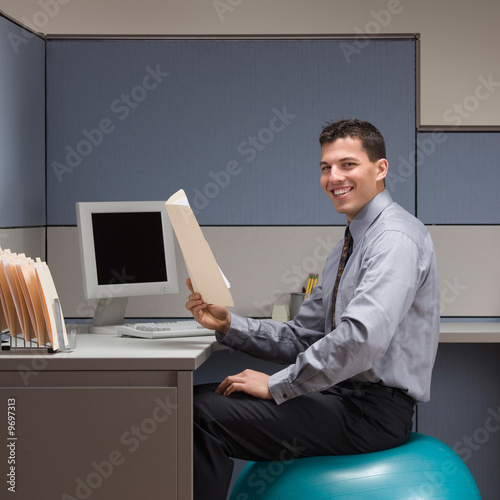 Photo: Smiling businessman sitting on exercise ball at desk in cubicle Smiling businessman sitting on exercise ball at desk in cubicle