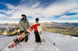 Two snowboarders stand on a peak over looking a spectacular view