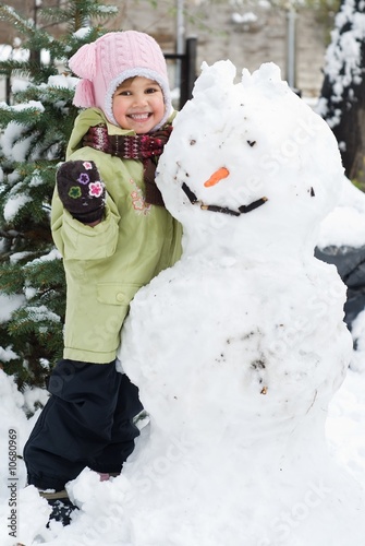 Photo: Cute little girl playing in winter backyard with a huge snowman Cute little girl playing in winter backyard with a huge snowman