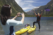 Woman taking picture of man hoisting oar of kayak over head on shore of mountain lake