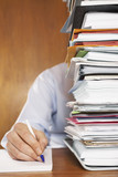 Photo: Man writing, close-up of arm and hand, sitting behind stack of paperwork at desk Man writing, close-up of arm and hand, sitting behind stack of paperwork at desk
