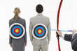Photo: Two business people with targets on backs while man aims bow and arrow close-up of hands, against white background Two business people with targets on backs while man aims bow and arrow close-up of hands, against white background