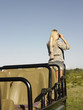 Photo: Young woman on safari, standing in jeep, looking through binoculars, back view Young woman on safari, standing in jeep, looking through binoculars, back view