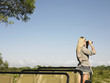 Photo: Young woman on safari, standing in jeep, looking through binoculars, back view Young woman on safari, standing in jeep, looking through binoculars, back view