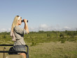 Photo: Young woman on safari, standing in jeep, looking through binoculars, side view Young woman on safari, standing in jeep, looking through binoculars, side view