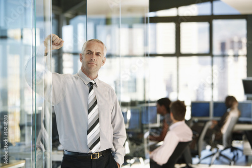 Photo: Businessman leaning on door in office, with office workers in background. Businessman leaning on door in office, with office workers in background.