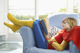 Photo: Young boy 7-9 sitting in armchair reading, wearing superhero costume, side view Young boy 7-9 sitting in armchair reading, wearing superhero costume, side view