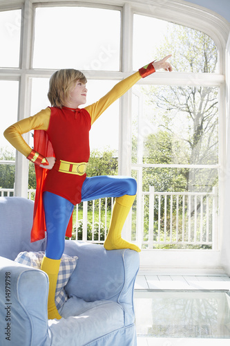 Photo: Young boy 7-9 wearing superman costume standing on armchair and pointing Young boy 7-9 wearing superman costume standing on armchair and pointing