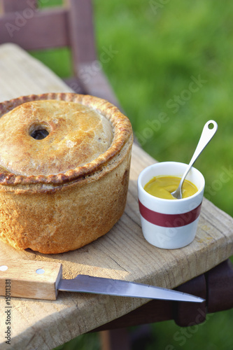 Photo: Pork pie, knife and cup of mustard on table outdoors Pork pie, knife and cup of mustard on table outdoors