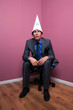 Businessman sat in corner wearing a dunce hat