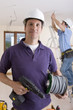 Photo: Electrician holding cable spool and drill with co-worker wiring ceiling in background Electrician holding cable spool and drill with co-worker wiring ceiling in background