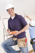 Smiling electrician holding voltmeter on ladder with co-worker wiring ceiling in background