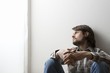 Young man in checked shirt sits with hands clasped against a wall