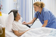 Female nurse using stethoscope on senior woman in hospital bed