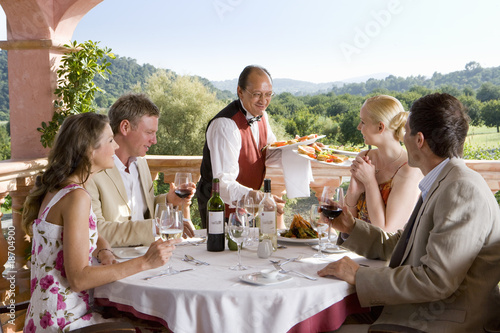 Photo: Waiter serving meals to well-dressed couples at table on restaurant balcony Waiter serving meals to well-dressed couples at table on restaurant balcony