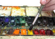 man's hand with paintbrush in over well-used watercolor paintbox