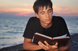 teenager boy reads book sitting on beach in evening