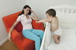 Young woman on arm chair holding her head and baby boy in crib