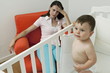 Young woman on arm chair holding her head and baby boy in crib
