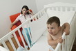 Young woman on arm chair holding her head and baby boy in crib