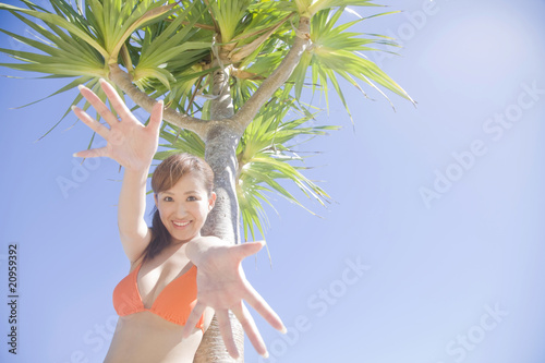 Foto: 椰子の木の側で手を伸ばす笑顔の水着女性 椰子の木の側で手を伸ばす笑顔の水着女性