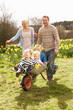 Father Giving Children Ride In Wheelbarrow Through Field Of Spri Father Giving Children Ride In Wheelbarrow Through Field Of Spri