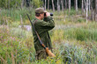 Hunter with rifle and binoculars in a forest`s swamp.
