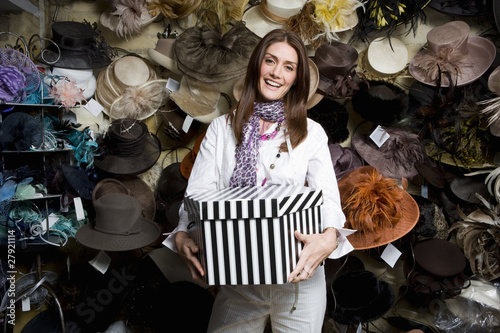 Photo: Saleswoman holding hat box in front of wall of ornate hats in shop Saleswoman holding hat box in front of wall of ornate hats in shop