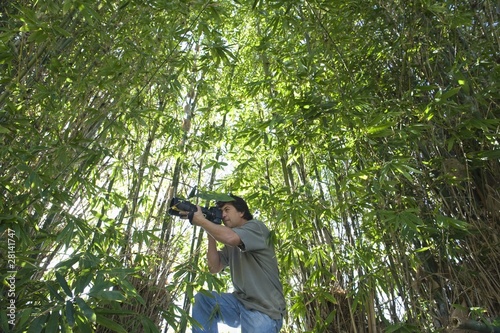 Photo: Photographer adjusts camera lens in bamboo forest, low angle view Photographer adjusts camera lens in bamboo forest, low angle view