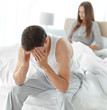 Photo: Stressed man sitting on his bed while his girlfriend working on Stressed man sitting on his bed while his girlfriend working on