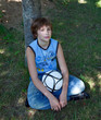 The teenager sits under a tree with a football.