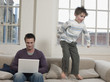 Father Using Laptop as Son Jumps on Sofa