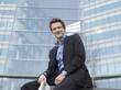 Mid-adult businessman sitting in front of office building, holding newspaper and smiling