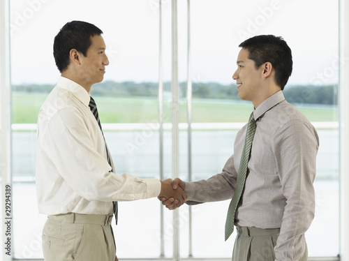 Photo: Businessmen standing, Shaking Hands in front of window, profile Businessmen standing, Shaking Hands in front of window, profile