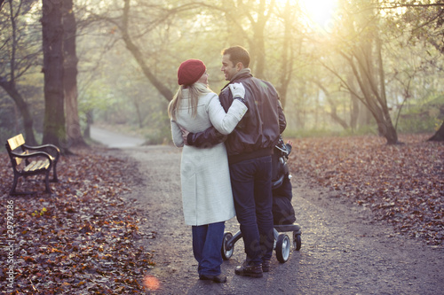 Photo: A young couple pushing a stroller in the park, rear view A young couple pushing a stroller in the park, rear view